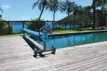 Pool mit Fitnessrolle auf Holzterrasse, umgeben von Palmen mit Blick auf Meer und Berge.